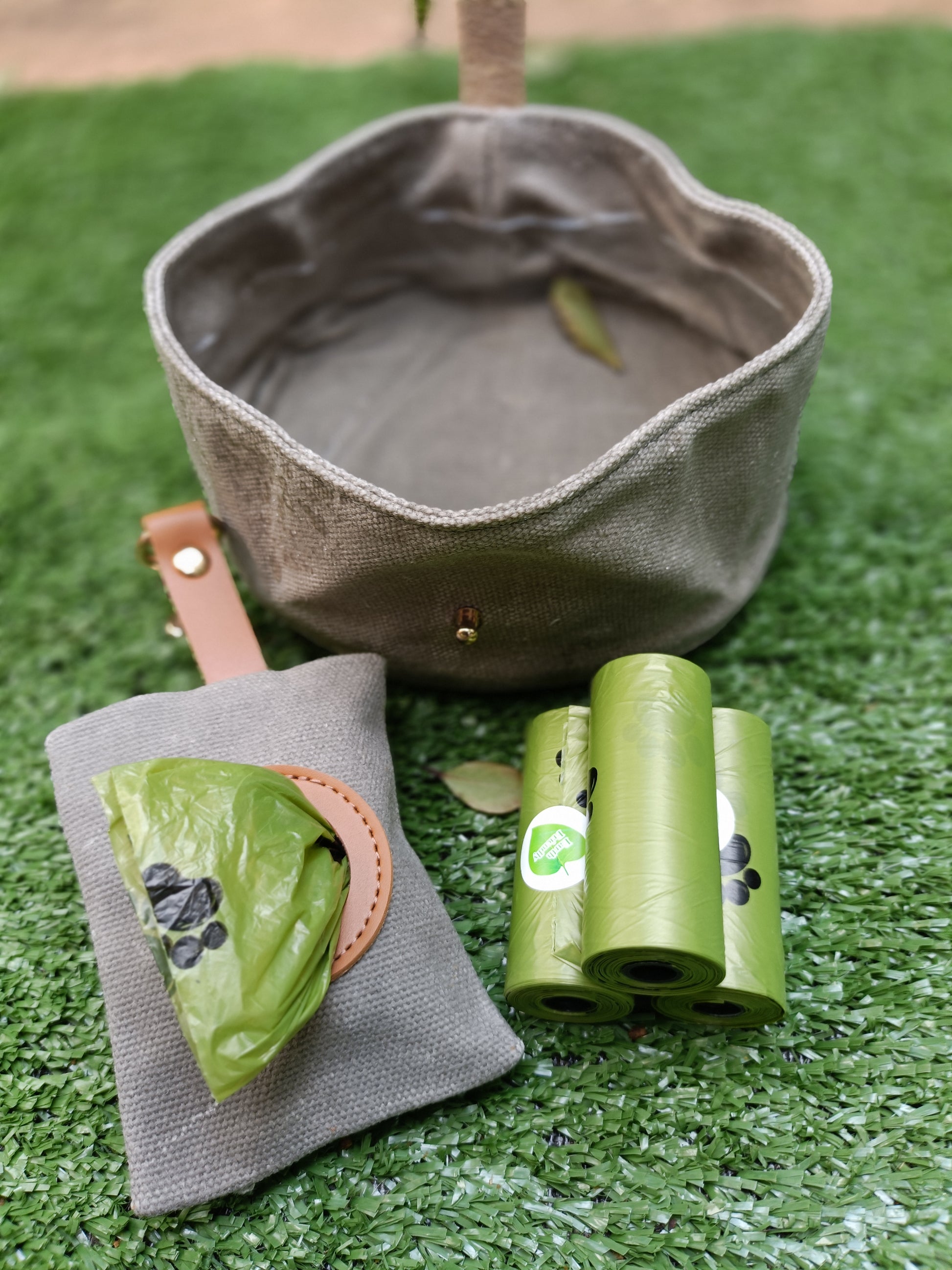 Grey canvas water bowl with three rolls of green waste bags on a grassy surface
